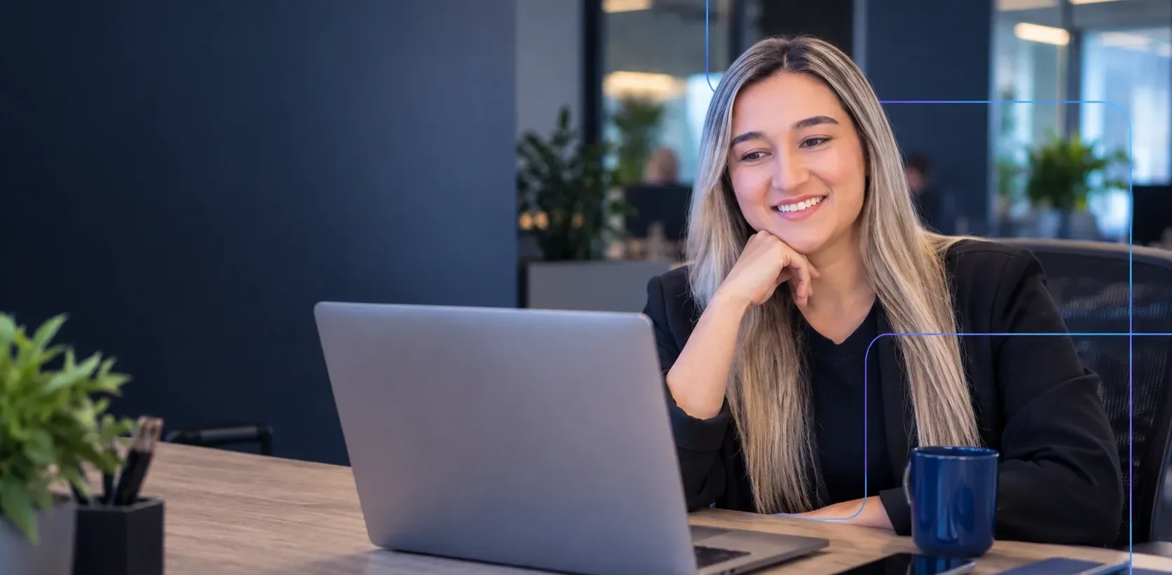 Mulher loira sorrindo em frente ao notebook em um escritório, com uma caneca azul ao lado